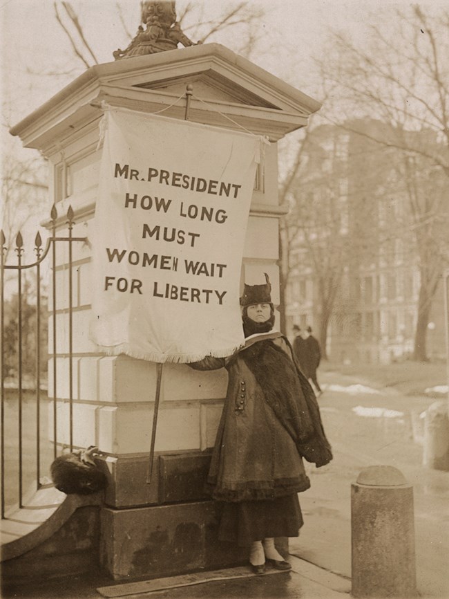 Woman standing next to a banner that reads, "MR. PRESIDENT HOW LONG MUST WOMEN WAIT FOR LIBERTY"