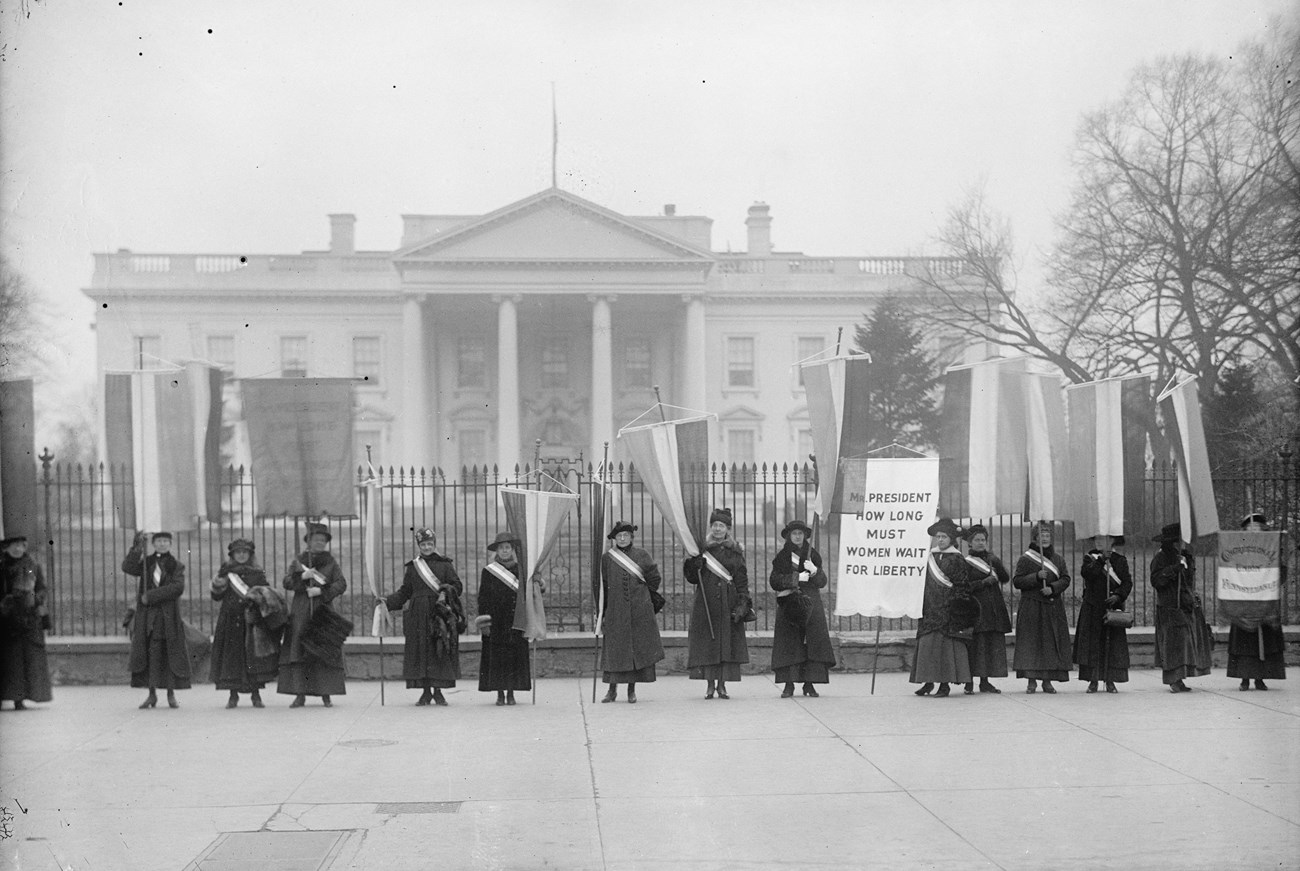 Group of women in long dresses holding banners outside of the White House.