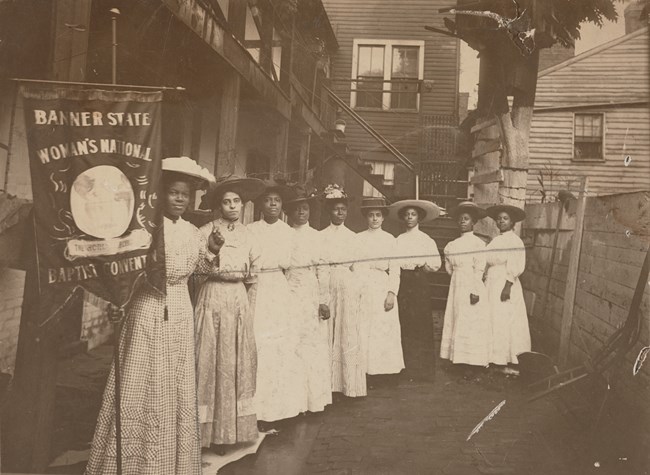 Nine African American women standing in a row holding a suffrage banner