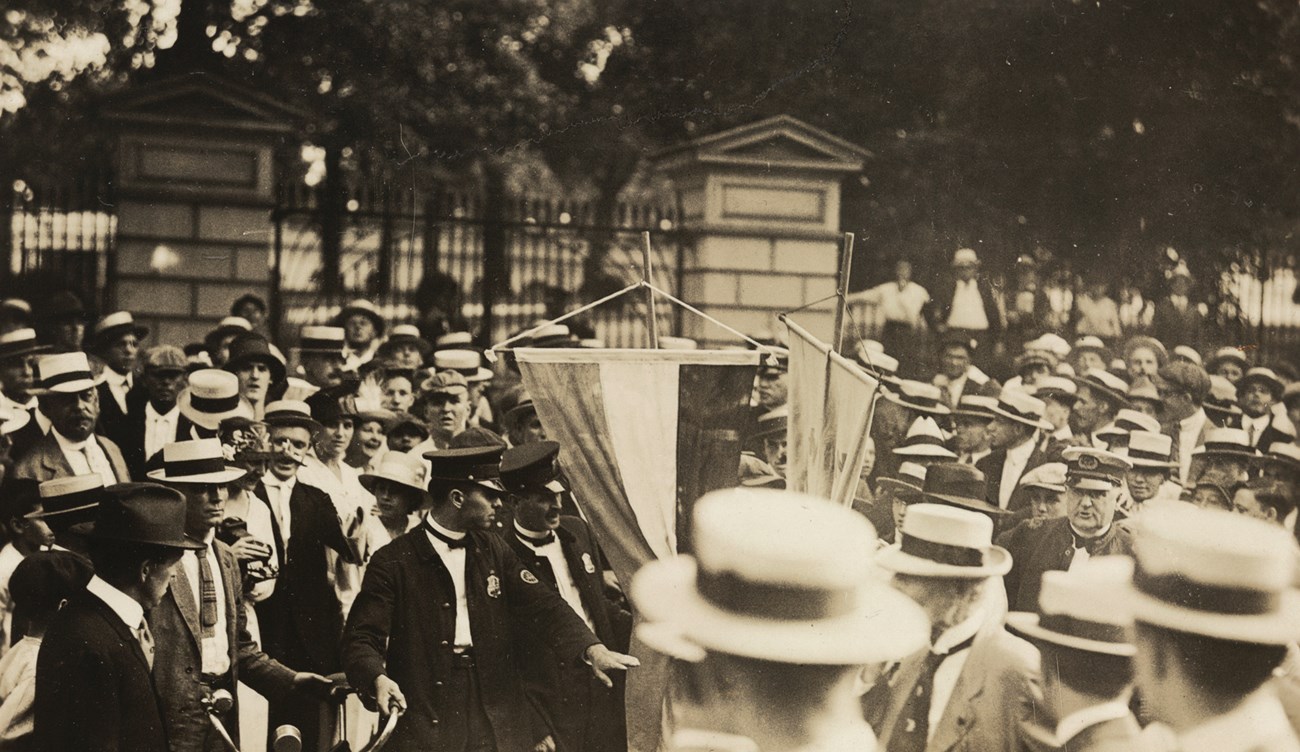 Two police officers arrest an unseen person in the middle of a large crowd of men