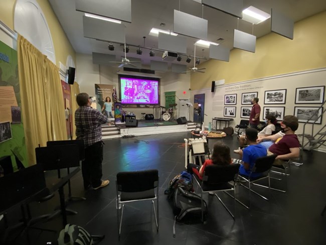 students seating in front of a small stage, watching a video on a big screen