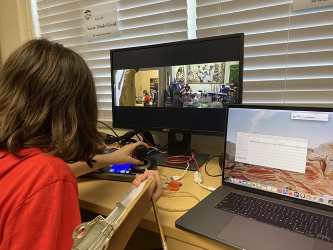 student in front of two computer monitors, working the mounted camera controller while other students are in front of the camera