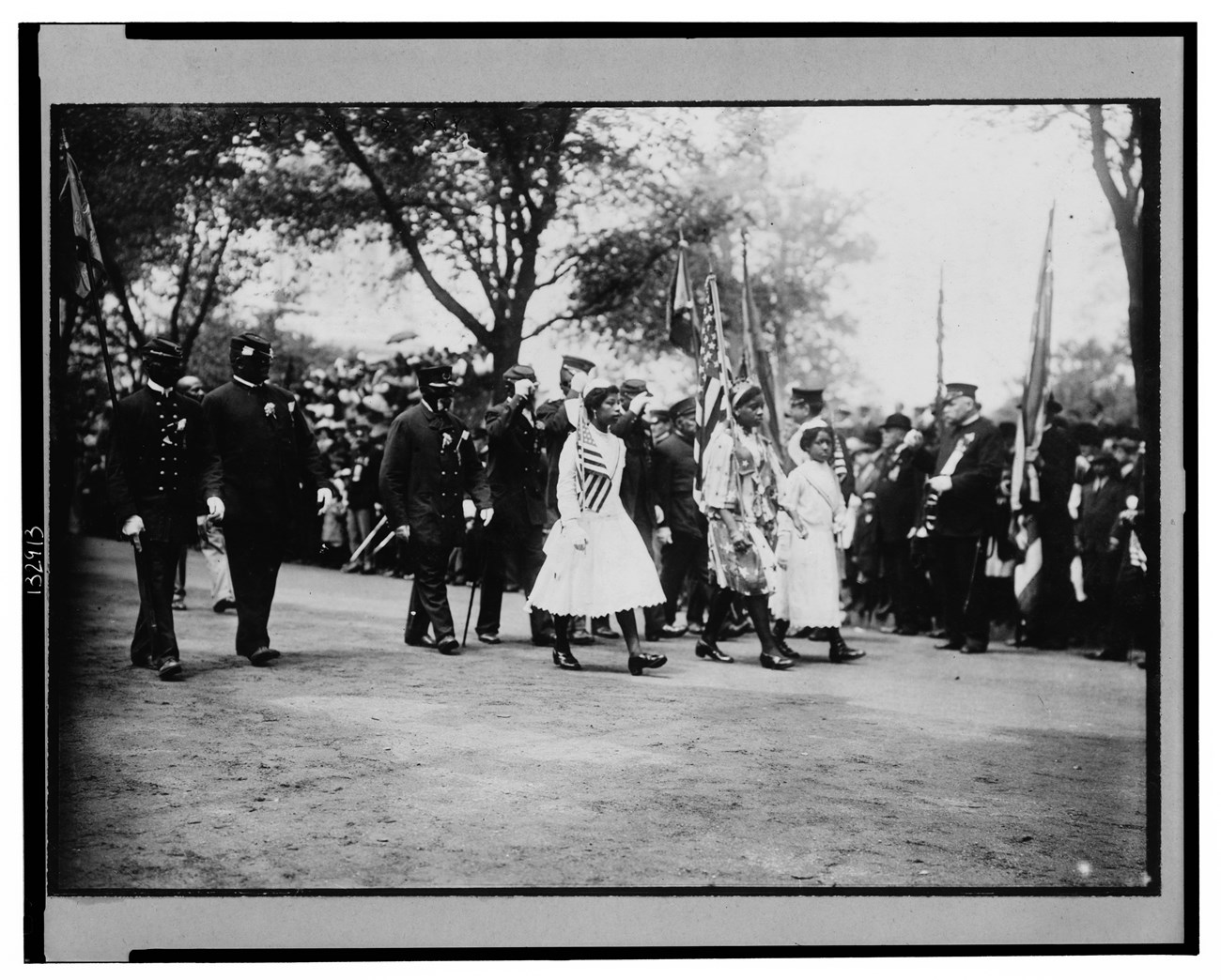 African American veterans marching in a New York City Memorial Day Parade, May 30, 1912. Marching in front of the veterans, 3 young Black women carrying American flags. In the background, a large interracial crowd of spectators.