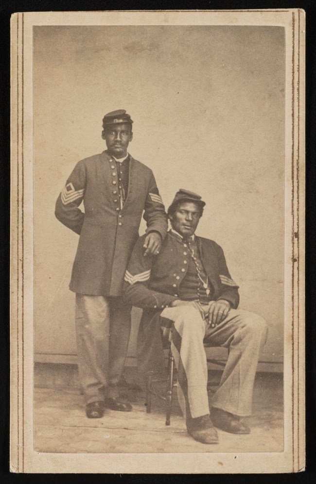 Two civil war era Black soldiers pose for their photo, one sits and the other stands.