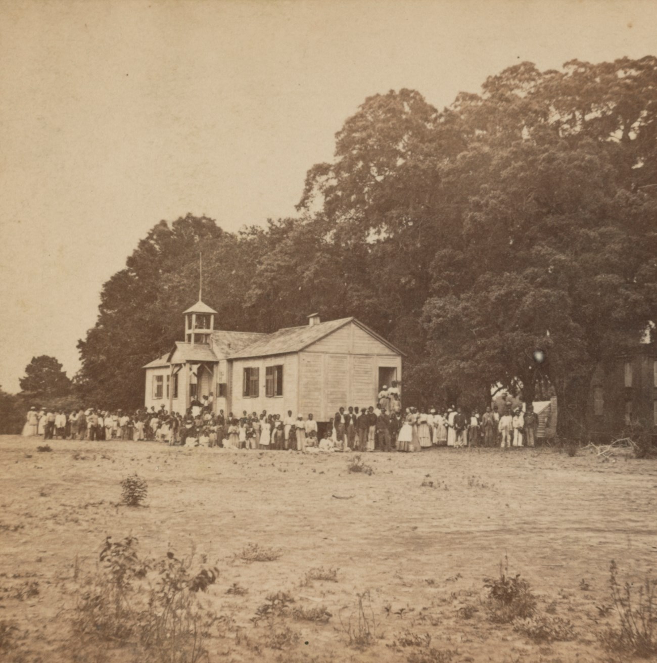 A small schoolhouse in front of a group of trees with students standing in front of the school.