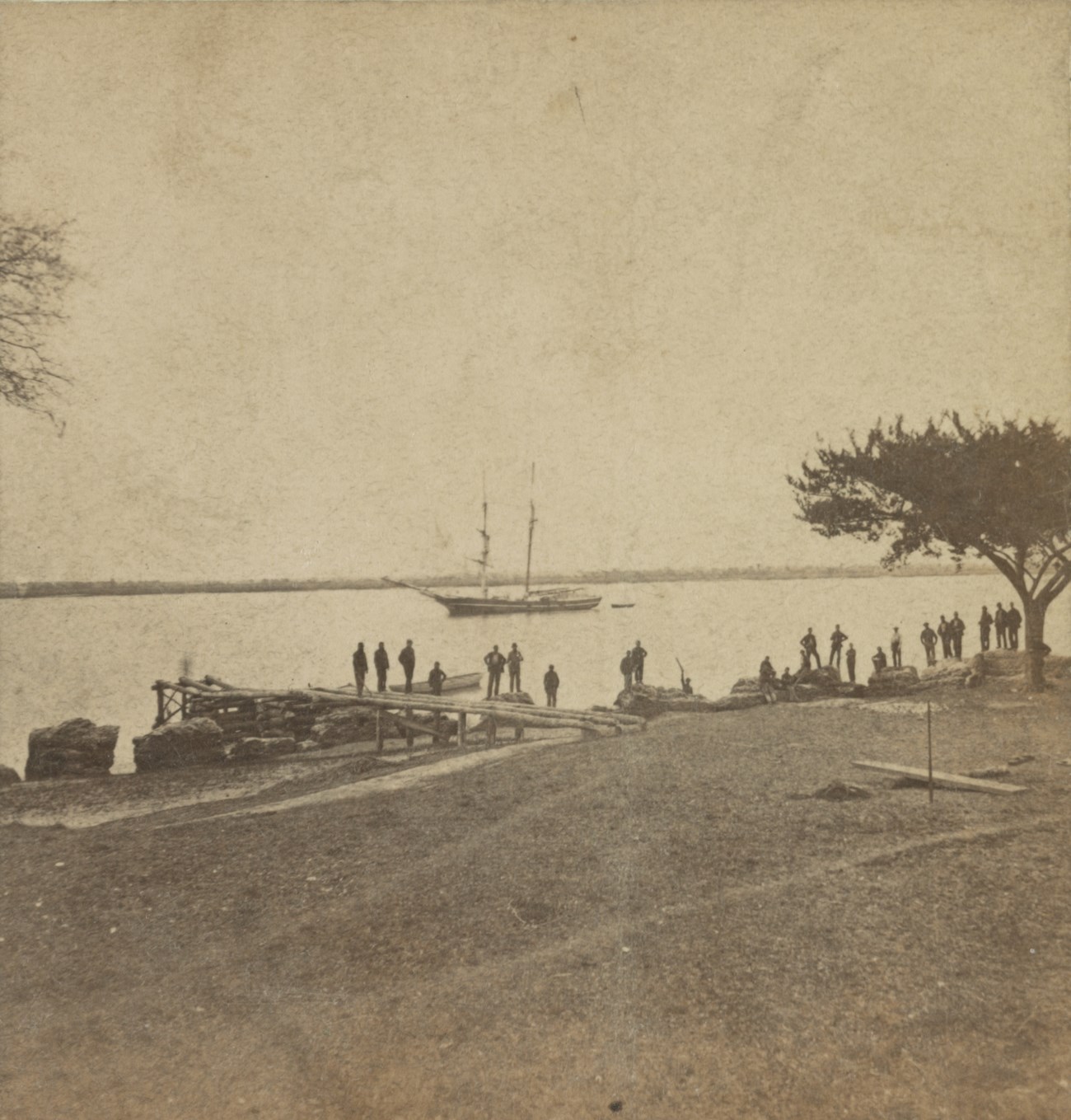 A group of men stand on the ruins of an old fort and a dock with water and a boat in the background.