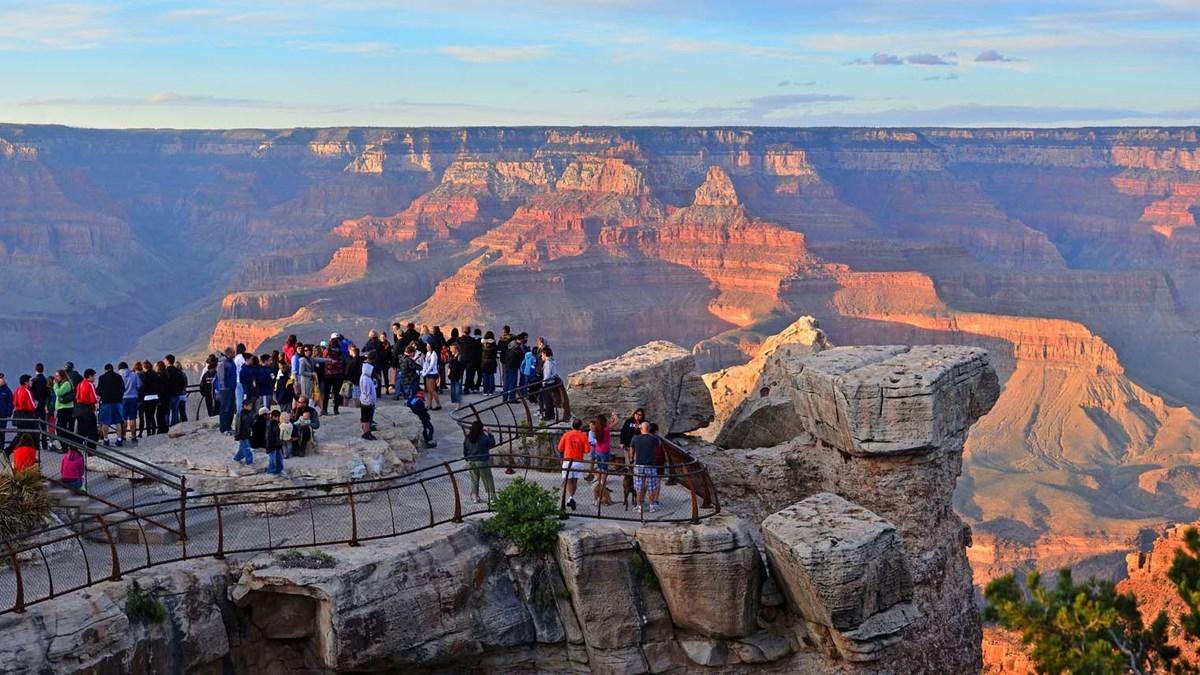 a crowd of sightseers behind railings at a scenic overlook. Distant peaks within a vast canyon are being illuminated by golden sunset light.