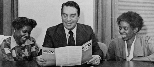 Two Black women sit on either side of a white man. They all look at a brochure he is holding.