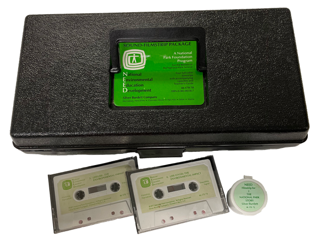 Plack plastic box with green label. Two audiocassettes and a filmstrip cannister beside it.