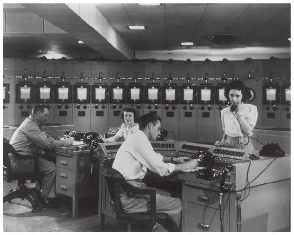 Two men work at desks while two women answer phones in front of filing cabinets in office setting