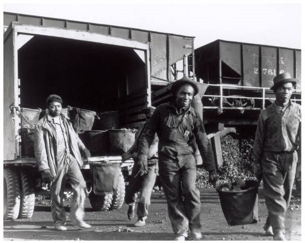 Four African American men carry large baskets away from a train with piles of coal