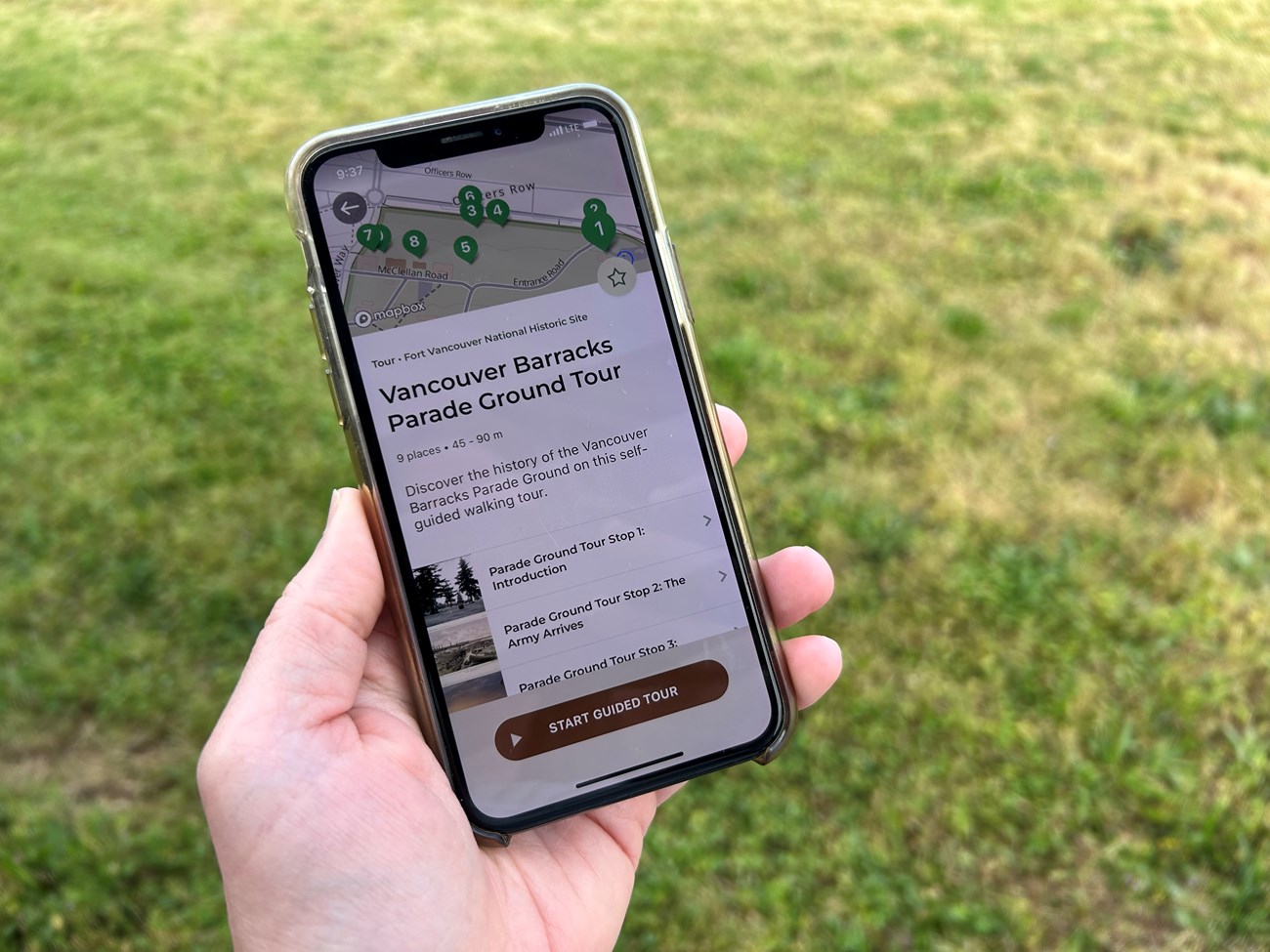 A hand is holding a smart phone, with the screen showing the first page of the Vancouver Barracks walking tour including text and a map with hotspots.