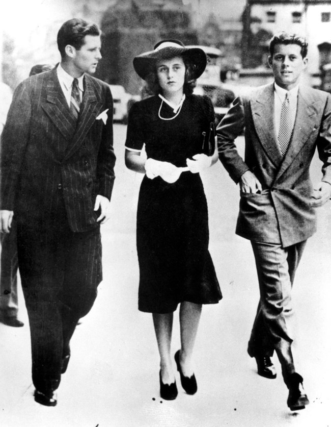 (L-R) Joe Jr., Kathleen, and John Kennedy walking in London.  The photo is black and white and the three are dressed in formal attire.