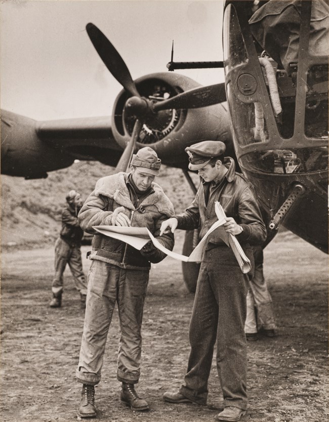 Two male pilots hold maps and charts in front of a plane.