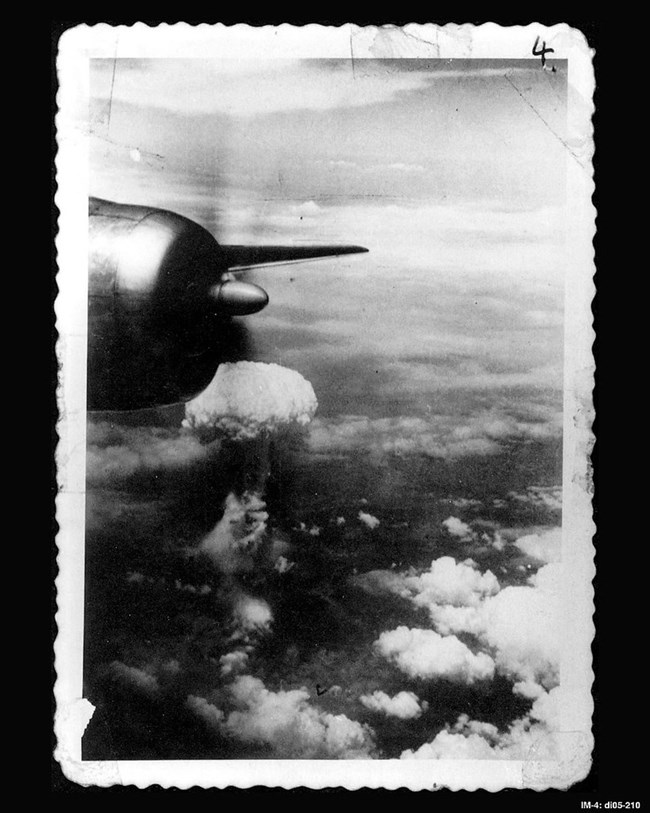 Black and white photo overlooking the wing of a plane, a mushroom cloud in the background