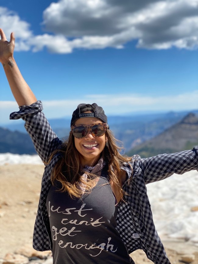 Woman stands on a mountaintop, smiling and raising her arms in celebration. Her t-shirt says “Just Can’t Get Enough.”