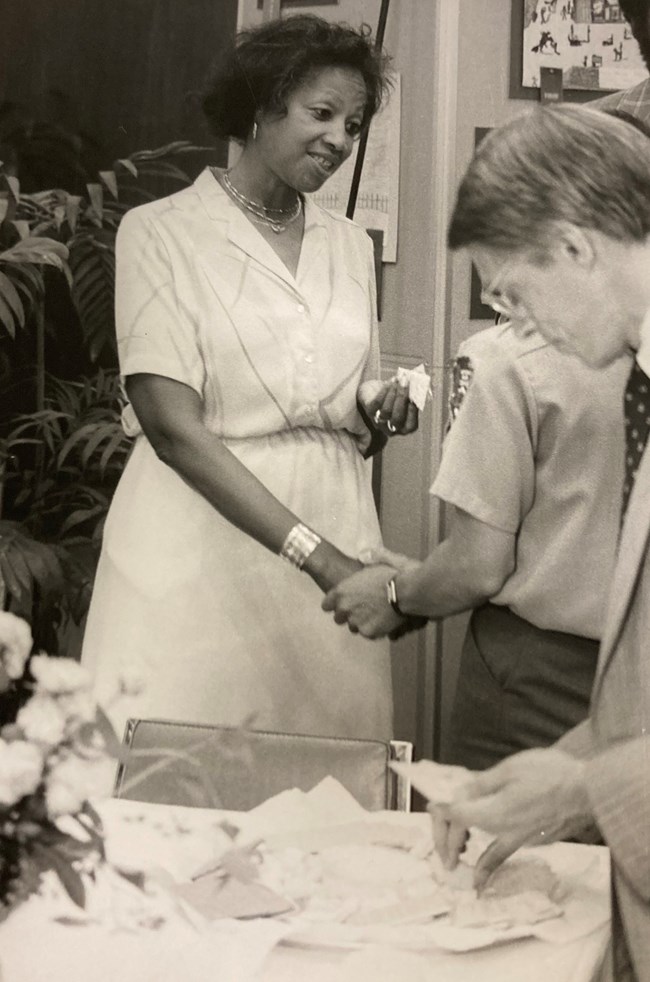 Dorothea Powell stands behind a table at a reception, wearing a dress and speaking with a ranger in NPS uniform who is clasping her right hand.