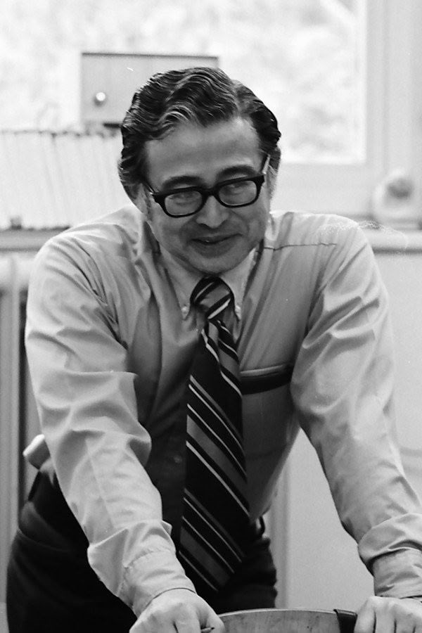 Middle-aged man wearing glasses and smiling while leaning on the back of a chair.