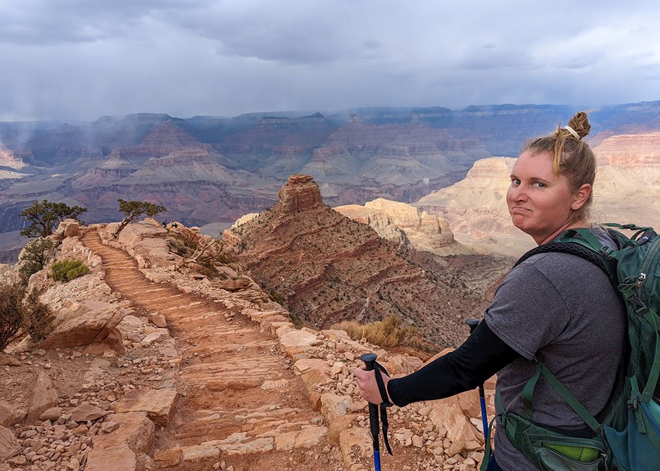 Holding hiking poles on a backcountry trail, a woman with her hair pulled up in a bun is looking at the camera and grimacing.
