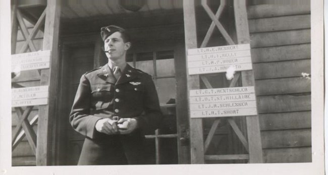 Smiling man in uniform, with pipe in his mouth, stands outside building with names of officers on signs beside him.