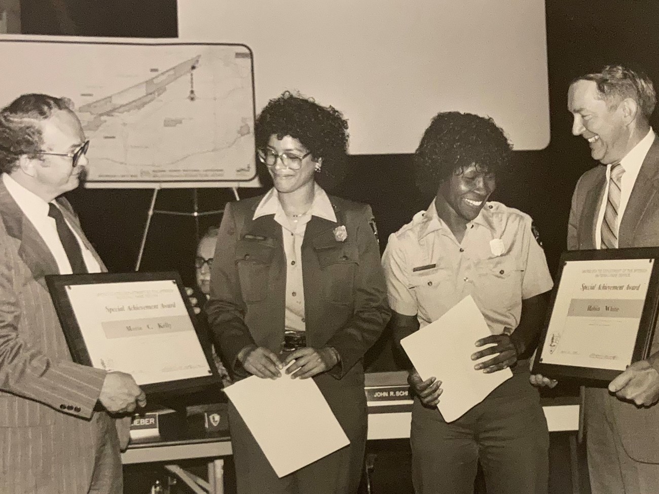 Marta Kelly and Robin White stand between two men in suits presenting plaques. The women wear NPS uniforms and hold papers in their hands.