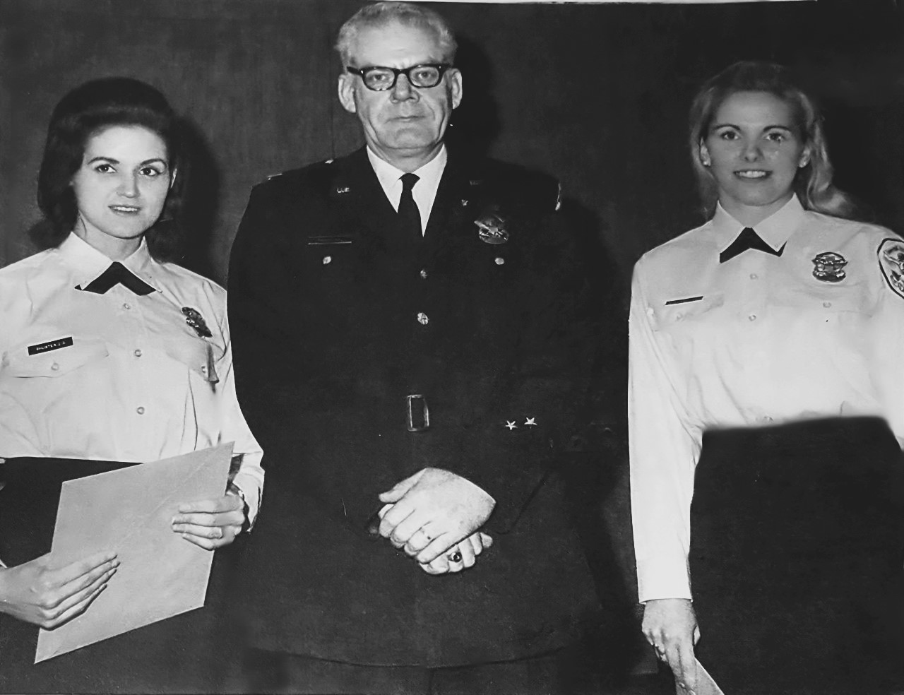 Two police women in uniform pose standing on either side of a policeman.