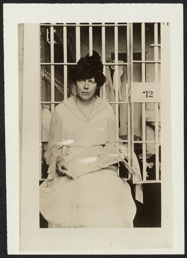 Informal portrait, Lucy Burns, three-quarter length, seated, facing forward, holding a newspaper in her lap in front of a prison cell, likely at Occoquan Workhouse in Virginia.