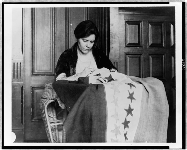 Alice Paul, half-length portrait, seated, facing slightly right, sewing suffrage flag.