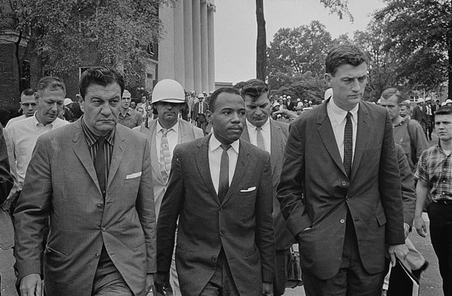 James Meredith stands between 2 US Marshalls. More marshals wearing helmets are seen in the crowd behind.