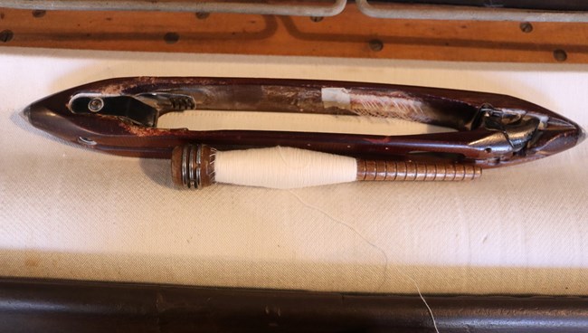 A hollow boat-like wooden vessel sits on a white piece of cloth. Next to it there is a skinny wooden dowel with groves, wrapped in white thread. At the end of the dowel there is a thicker piece of wood with metal strips.