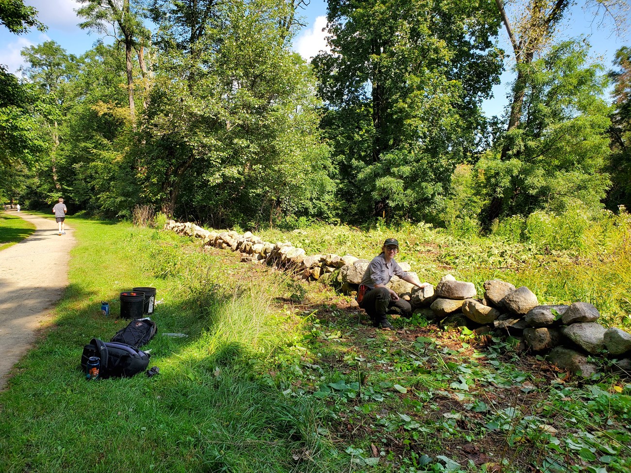 Woman in an NPS uniform kneels next to a stone wall. On one side of the wall is a grove of trees. On the other is a grass-bordered dirt path with a runner in the distance.