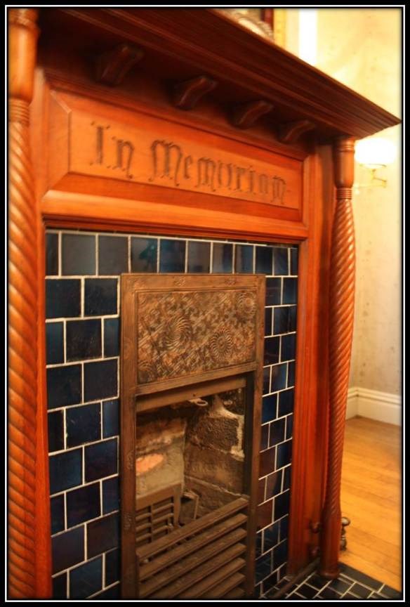 a fireplace that has wooden around the outside as welll as blue tiles. "In Memorium" is inscribed in the tile