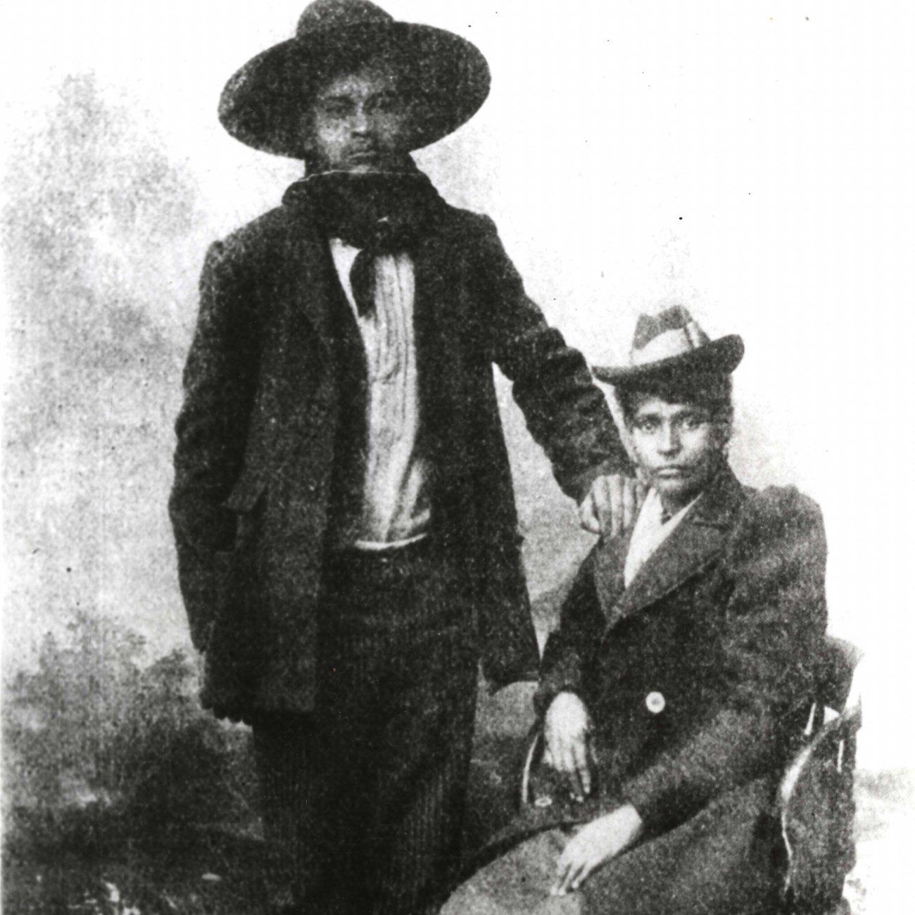 Black and white historic image of Cherokee Bill standing wearing a dark wide-brim hat with his hand on the shoulder of the woman sitting next to him.