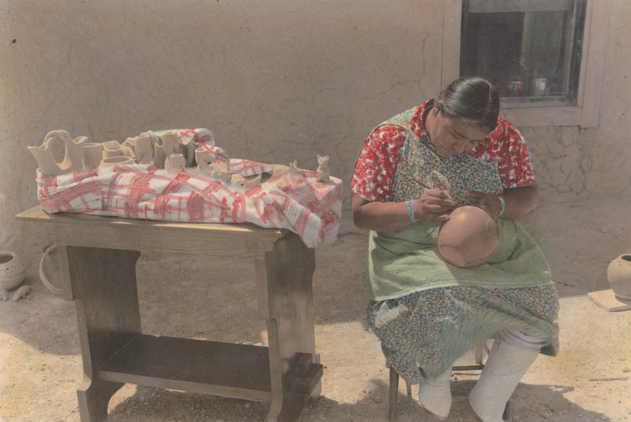 Legoria Tafoya sits outside using a paintbrush to paint a pot.