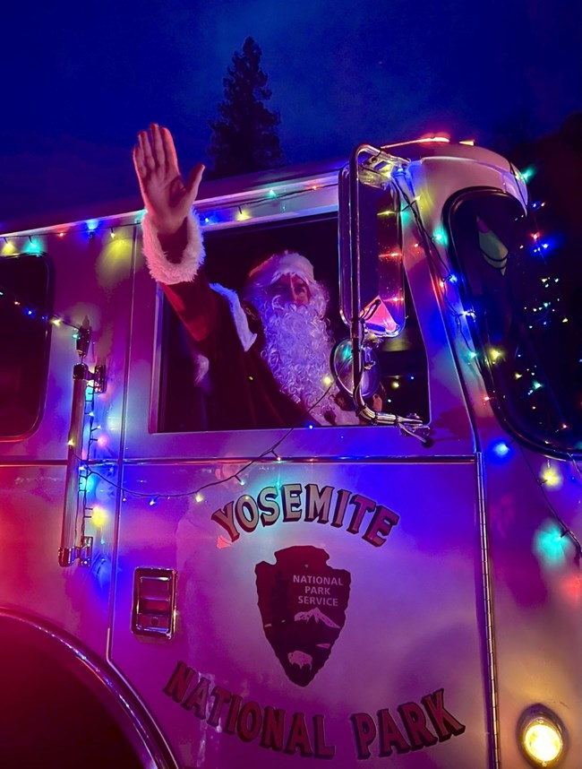 A man dressed as Santa waves from a fire engine decorated with multi-colored lights.