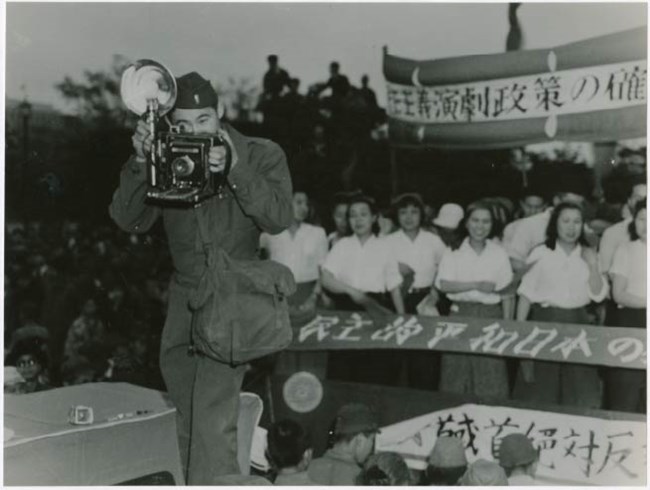A man standing in front of a crowd, taking a photo with a 1940s era camera.