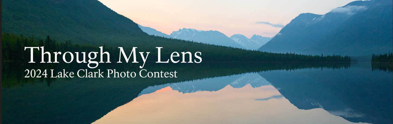 A lake reflects a sunset background with mountains and trees in the distance. In the center left reads white text saying "Through My Lens 2024 Lake Clark Photo Contest