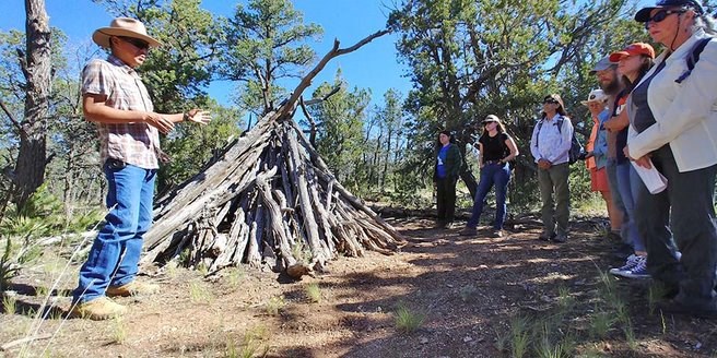 A man talks to a group of people arranged in a half circle, while he stands next to a structure made out of tree limbs.