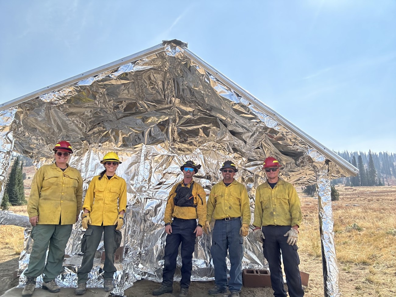 Five people in hard hats and fire gear standing in front of a house wrapped in silver foil.