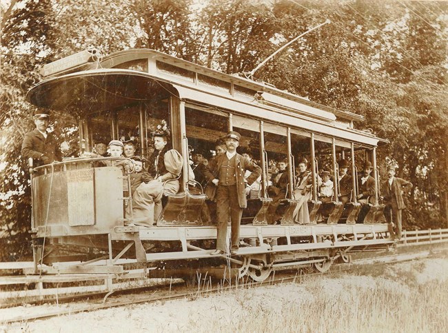 A group of people sit on a trolley as a conductor poses with hand on hips on the side