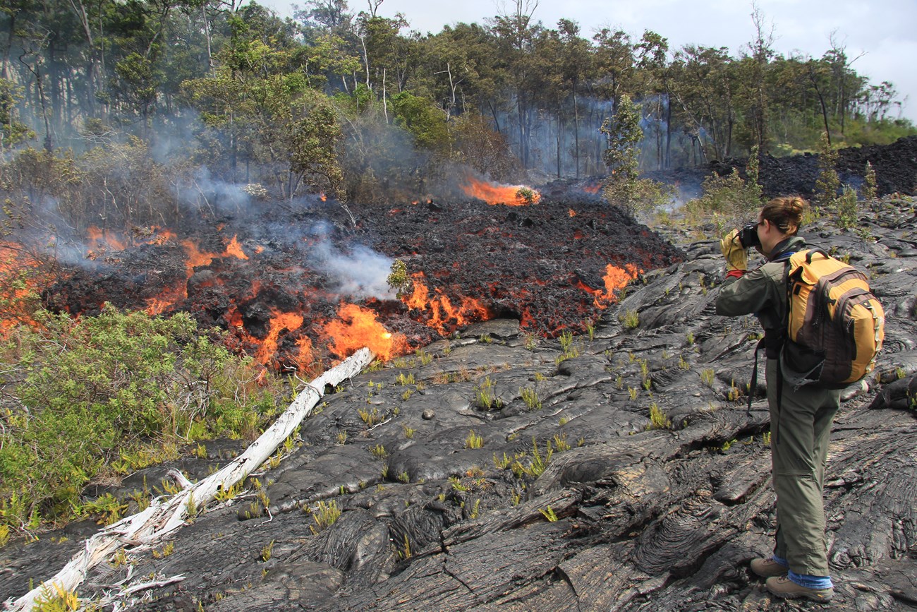 Photo of a person standing and watching molten lava flow across an older lava rock surface.