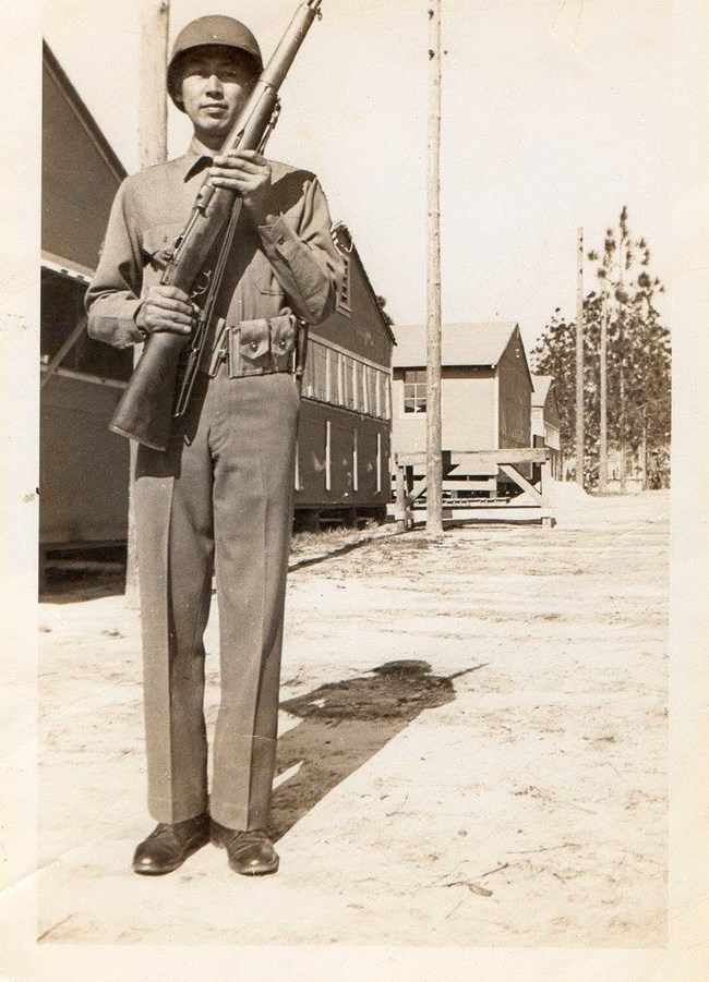 A man in an Army uniform stands with his firearm in front of a barrack.