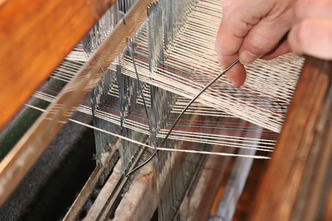 Close up shot of a hand holding a thin metal instrument which reaches through a hole in a thin strip of metal to draw a thread through