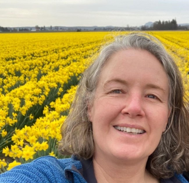 woman smiles at the camera with a field of bright yellow flowers behind her