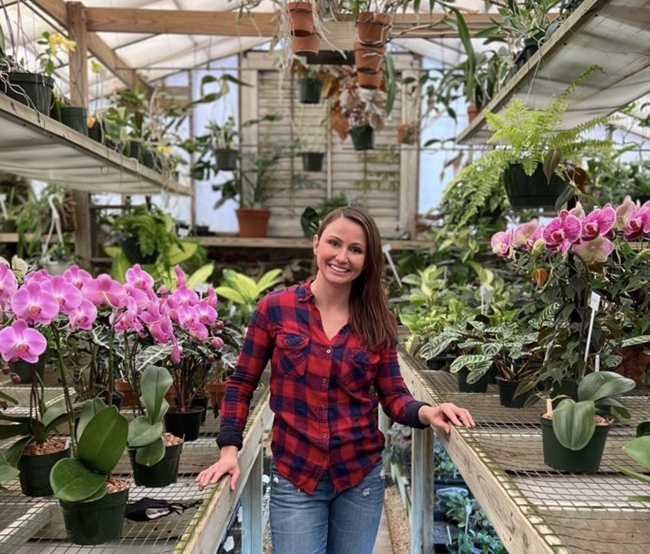 single woman stands in greenhouse between flowering plants in pots