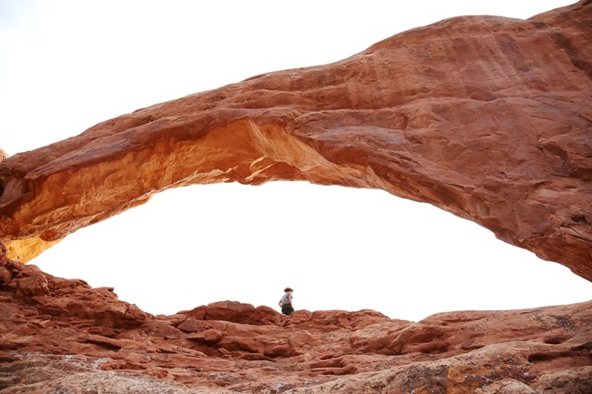 A park ranger in uniform stands inside an orange colored natural stone opening. The sky behind is white.