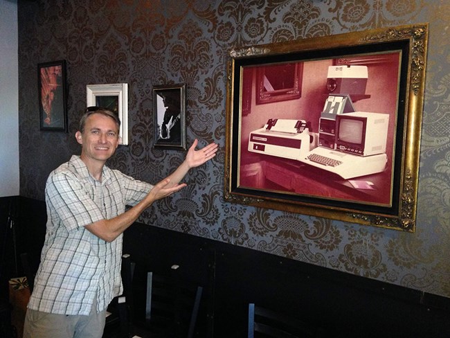 Person gestures excitedly to a framed picture of an old computer set up.