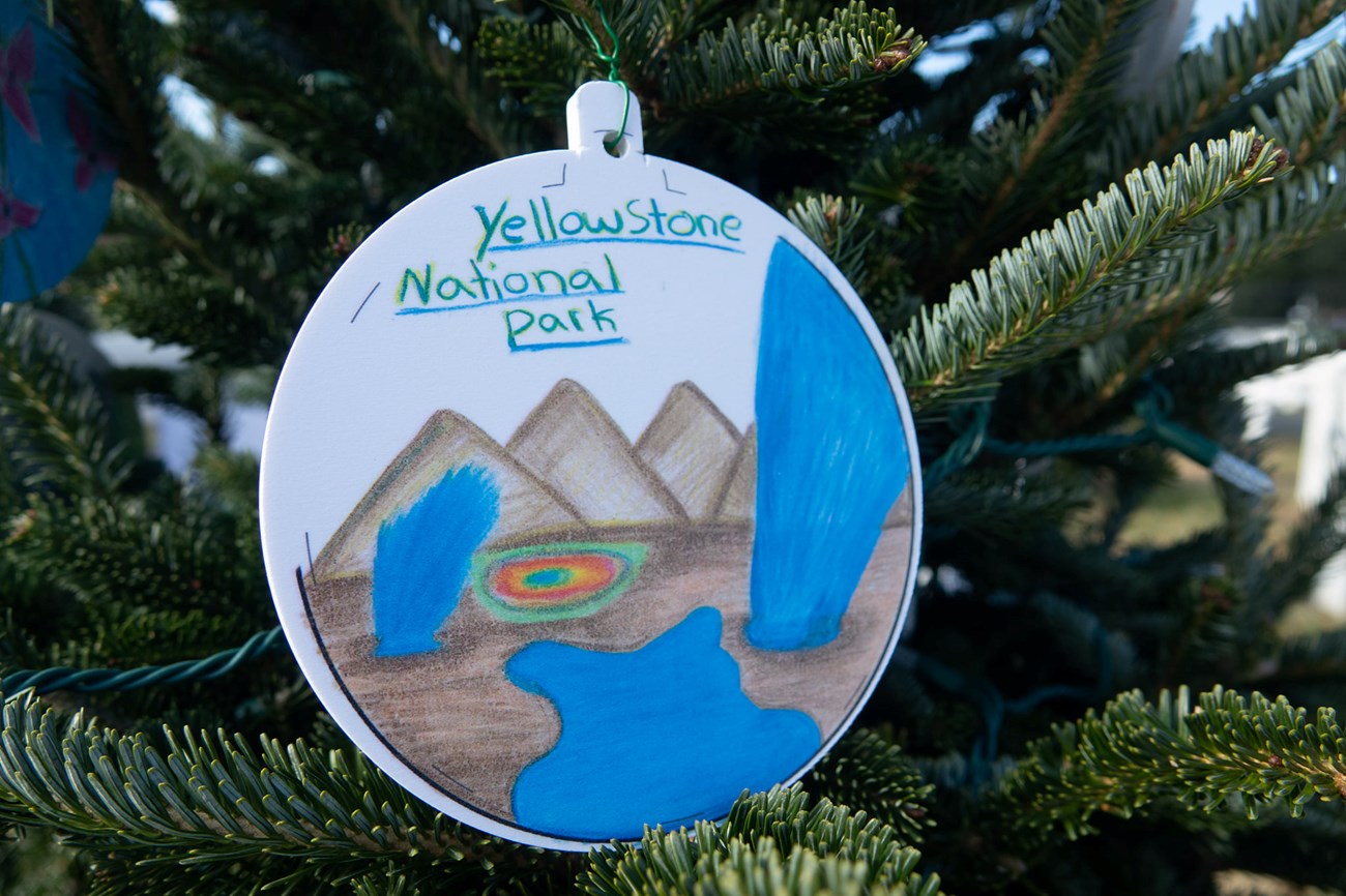 ornament with a drawing of mountains and yellowstone's geyser in the middle. Ornament is hanging from christmas tree