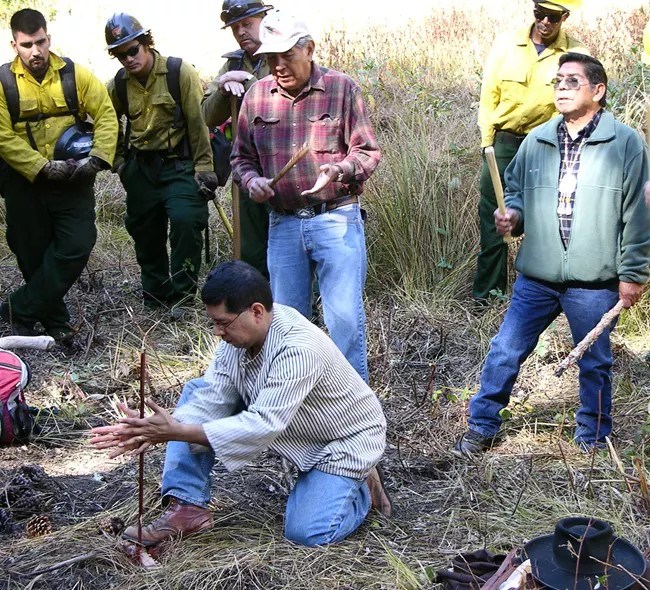 A man uses a stick to start a fire in a grassland ecosystem, with others watching. Some of the people watching are wearing Yosemite Fire uniforms.