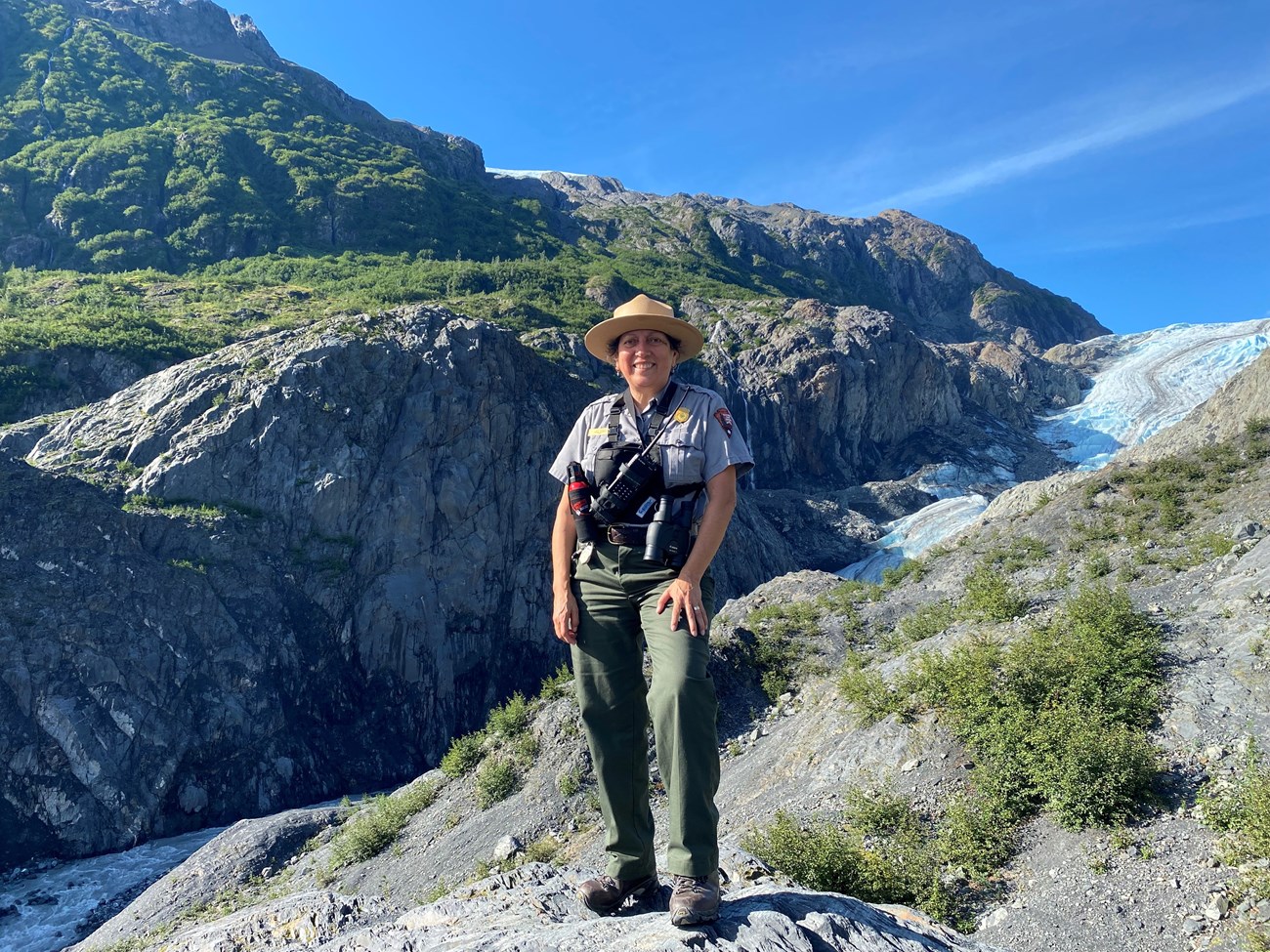 A ranger on a rocky terrain with a glacier coming down a valley.